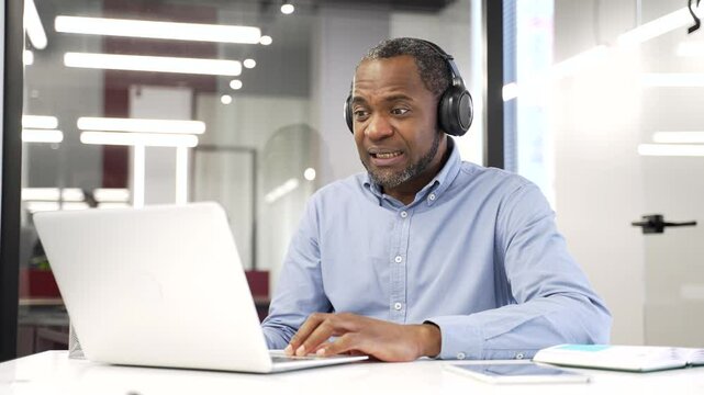 African american businessman in wireless headphones watching sports match or competition using laptop sitting in office. Happy man cheering for bids at auction, celebrating success showing achievement