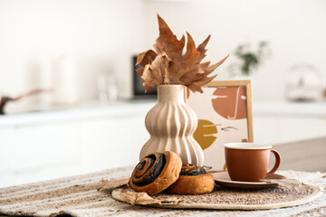 Wicker mat with cup of hot tea, tasty poppy buns and autumn leaves on table at home