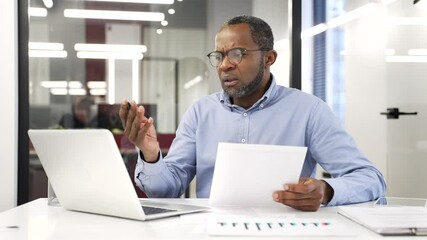 Disappointed african american businessman having difficulty with paperwork sitting at a desk in business office. Puzzled сonfused financier looks at documents and laptop and cannot understand problem