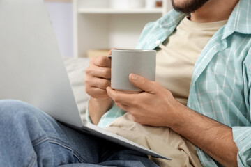Young man with ceramic cup and laptop sitting at home, closeup