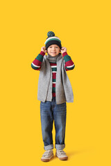 Cute little boy in warm sweater with scarf and winter hat on yellow background