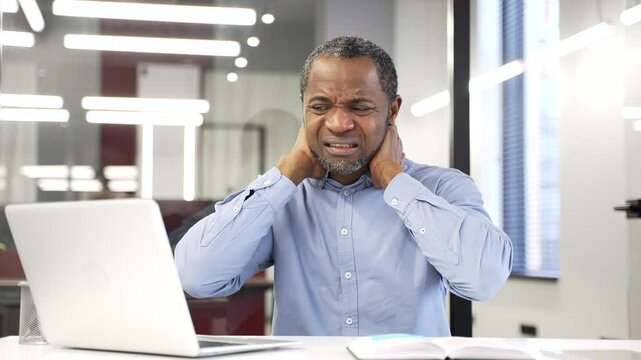 Tired american businessman businessman suffering from neck pain while working on laptop sitting at desk in business office. Upset black man feels discomfort, massages and rubs sore muscles, stretches
