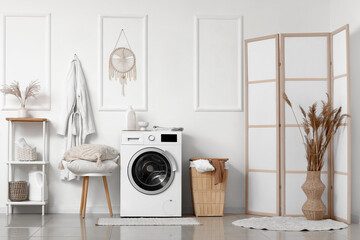 Modern washing machine with laundry basket and pillows on stool near white wall in bathroom
