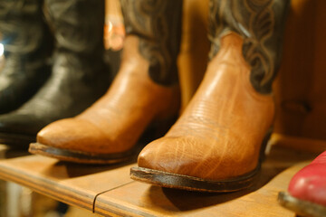 Close-up of tan leather cowboy boots with intricate stitching on wooden shelves, showing wear. Darker boots in the background add depth. Warm lighting highlights the rustic, vintage texture.