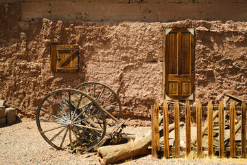 A rustic scene features an adobe wall with wooden door and window, a weathered wagon wheel, and a broken fence in a desert setting