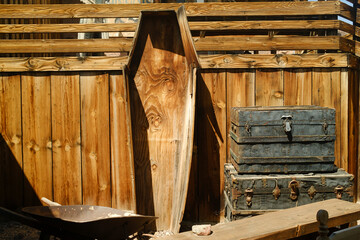 A weathered wooden coffin leans against a rustic fence, next to old metal trunks and a wheelbarrow