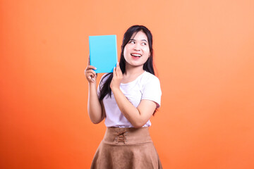 Naklejka premium A woman in a white shirt and brown skirt holds a blue book while looking off to the side with orange background