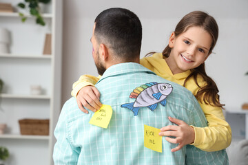 Happy little daughter attaching paper fish and sheets with text KICK ME to her father's back at...