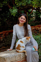 A woman in a striped dress sits on a stone ledge with a white and orange cat beside her; she holds...