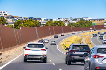 A multi-lane highway or major road with cars, bordered by tall sound barrier walls. A modern residential neighborhood with suburban houses or real estate development on a hillside in Australia