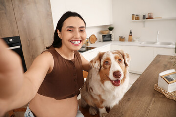 Young happy pregnant woman with cute Australian Shepherd dog taking selfie in kitchen at home