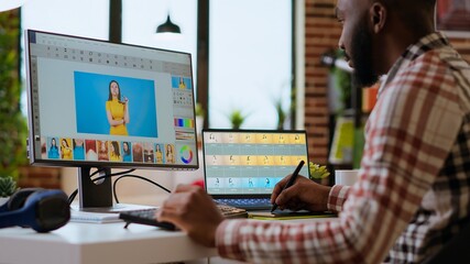 Male african american designer works with photo editing software in his creative workspace at home. Using dual monitors to ensure productivity and innovation in the graphic design world. Camera B.