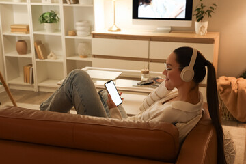 Young woman in headphones with mobile phone lying on sofa at home in evening