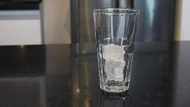 Cubos de hielo cayendo en un vaso de vidrio transparente con gotas de agua en una cocina.