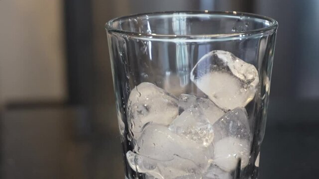 Cubos de hielo cayendo en un vaso de vidrio transparente con gotas de agua en una cocina.