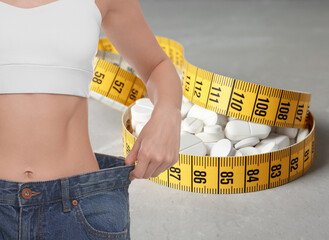 Weight loss. Slim woman in big jeans, measuring tape and pills on grey background, closeup