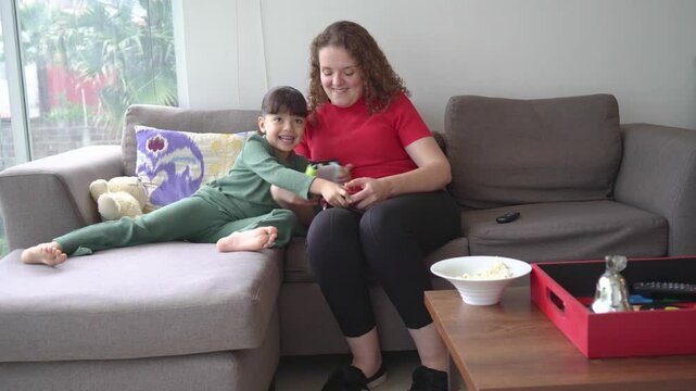 A mother and daughter play video games together in the living room, trying to outsmart each other to win. they laugh and tease each other while enjoying their competitive moment.