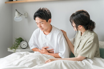 朝寝室で腹痛に悩む男性と心配する女性・夫婦・カップル
