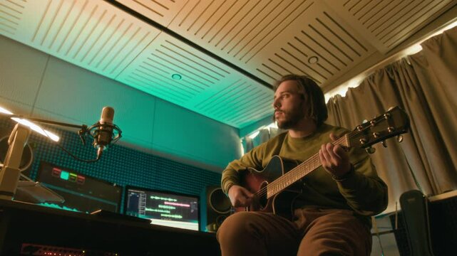 Low angle shot of professional musician or artist playing upbeat melody on guitar in modern neon lit recording studio. Recording new song or podcast, audio sequences on computer screens in background