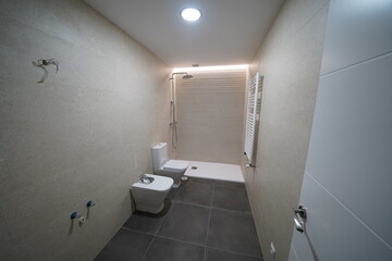 Modern bathroom under renovation with toilet, bidet, wall radiator, and installed shower. LED ceiling lights. Missing sink, mirror, and shower screen. Minimalist style with neutral tones.