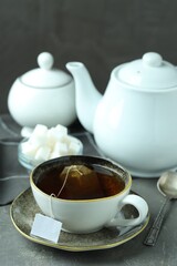 Tea bag in cup with hot drink, teapot, sugar cubes and spoon on gray textured table
