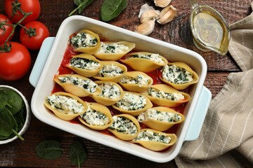 Delicious conchiglie pasta with ricotta cheese, spinach and tomato sauce in baking dish on wooden table, flat lay