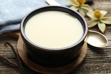 Tasty condensed milk, spoon, vanilla pods and flowers on wooden table, closeup