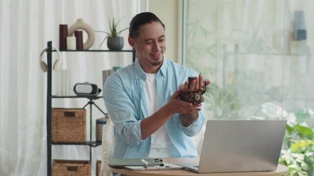 Medium copy space shot of happy multicultural owner calming domestic python snake while sitting at table in front of laptop at home office