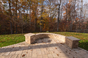 A serene backyard with a curved stone wall and patio pavers, surrounded by bare trees, autumn foliage, and scattered leaves.