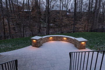 A peaceful backyard patio covered in fresh snow, featuring a curved stone wall with glowing lights. Black railings and bare trees enhance the serene winter ambiance at dusk.