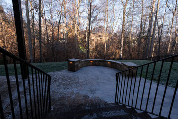 A backyard patio lightly frosted on a crisp winter morning, featuring a curved stone wall with soft glowing lights. Bare trees and distant homes add to the tranquil atmosphere.