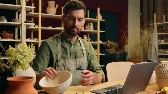 Happy joyful Caucasian man businessman male guy small business owner entrepreneur craftsman potter typing laptop online checking product porcelain crockery bowl design computer work in pottery store