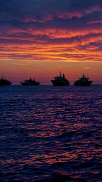Wide Shot of Sunset over the horizon with silhouettes of a fleet of ships, fiery orange and deep blue tones, warm, fading light, mood of awe and timeless beauty. Vertical video backgrounds
