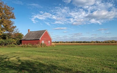 Fototapeta premium Charming red barn stands alone in a vast field under a bright blue sky with fluffy clouds
