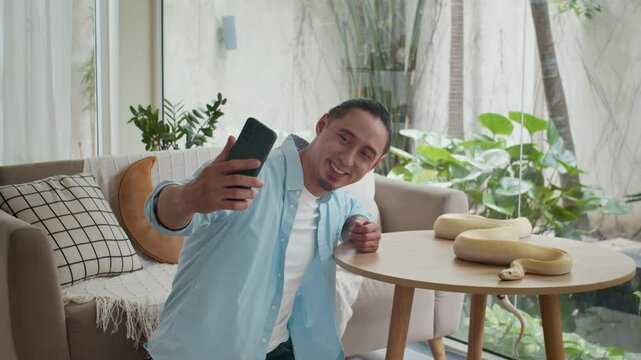 Wide shot of multi-ethnic man taking selfies on mobile phone with albino python snake resting on table at home