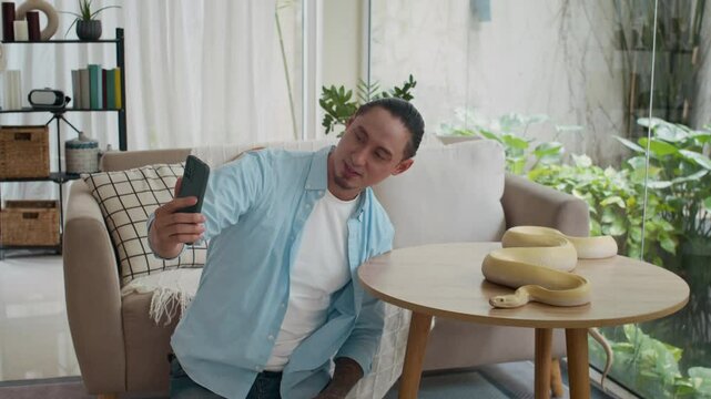 Wide shot of multicultural male owner talking on camera of smartphone about albino python snake wriggling on table in living room
