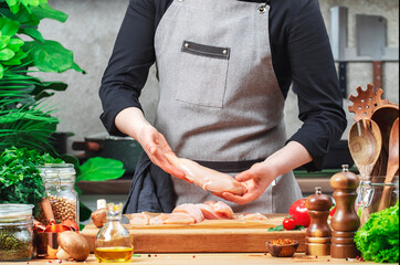 Chef in apron holding raw chicken breast fillet. Cozy kitchen with wooden table, kitchenware, vegetables, herbs and ingredients for cooking. Healthy eating, culinary, recipes, food blogging