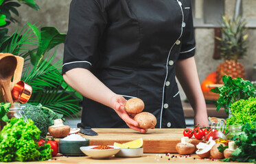 Chef in uniform holds fresh organic brown mushrooms. Cozy kitchen with wooden table, kitchenware, vegetables, herbs and ingredients for cooking. Healthy vegan food, culinary, recipes, food blogging