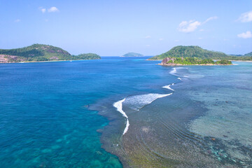 Drone shot of port glaud beach, island in the foreground, coral reef barrier, granite stones, white sandy beach and clear turquoise water, Mahe, Seychelles.j