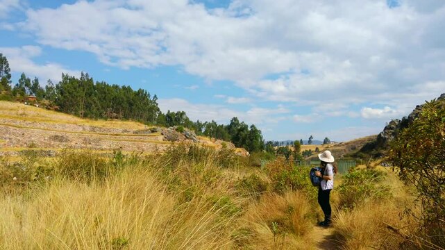 Woman tourists with Peruvian hat and backpack visiting the terraced ruins of Inkilltambo. Wide shot, pan right panoramic view