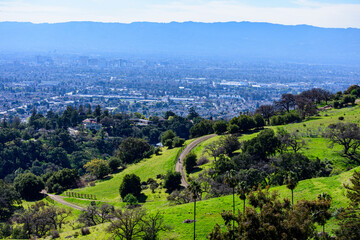 A winding dirt trail bordered by fencing cuts through green hills with scattered trees, overlooking...