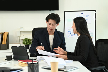 Two people in suits are talking at a desk with a whiteboard behind them. One of them is writing on a piece of paper.