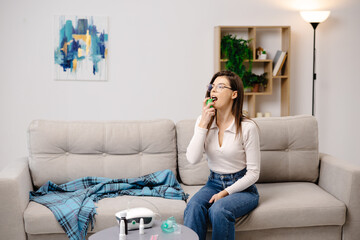 Obraz premium Young woman wearing casual clothing, using an analgesic spray to soften the throat, sitting on a sofa in a home interior
