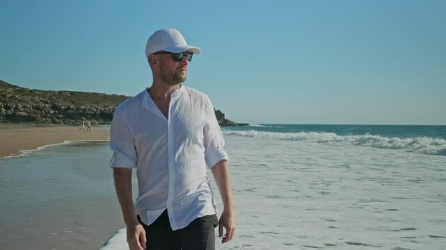 Middle aged Male model walking along the sandy coast on summer. Man  walks alone on a sandy beach. Adult man walking on a sandy beach against background of the ocean on summer sunny day, slow motion.