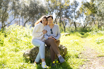 Fototapeta premium Adult daughter hugging senior mother sitting on rock in park