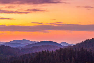 Colorful mountain sunrise over a dense forest. A breathtaking sunrise paints the sky in vibrant hues of orange, pink, and yellow over a range of mountains covered with dark evergreen forests.