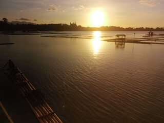 Floating fish cages on the lake are illuminated by the golden sunlight during sunset with a huge tree in the foreground. Silhouettes