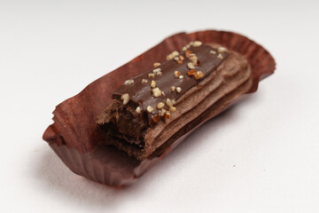 A bitten chocolate éclair showing its creamy filling, garnished with chopped nuts on top, served in a brown paper liner.