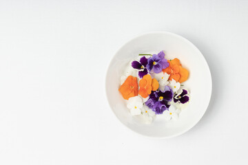 A top-down view of a white bowl with edible flowers in orange, purple, and white, arranged...