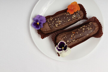 Two chocolate ?clairs in dark brown wrappers, topped with nuts and edible flowers, arranged on a white plate with a clean, minimalist background.